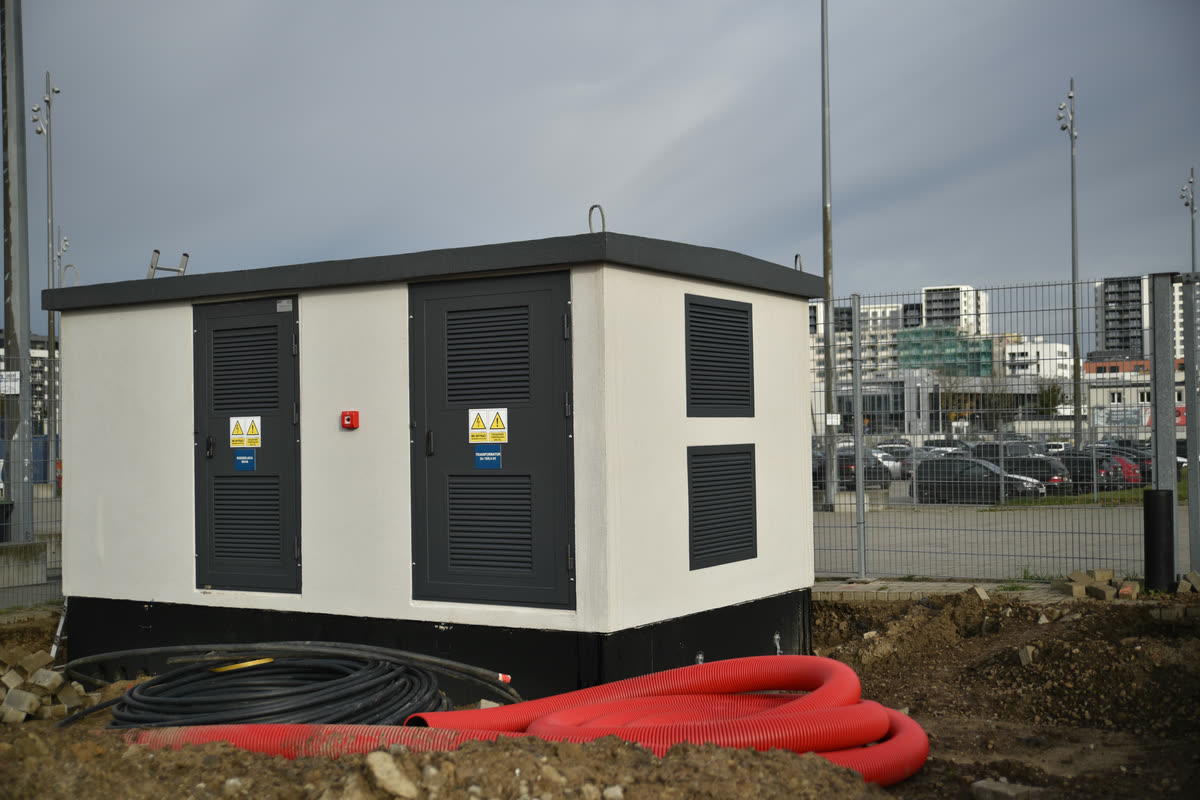 Prefabricated compact substation in a light concrete enclosure with dark ventilated doors, set on site with red cable ducts in the foreground