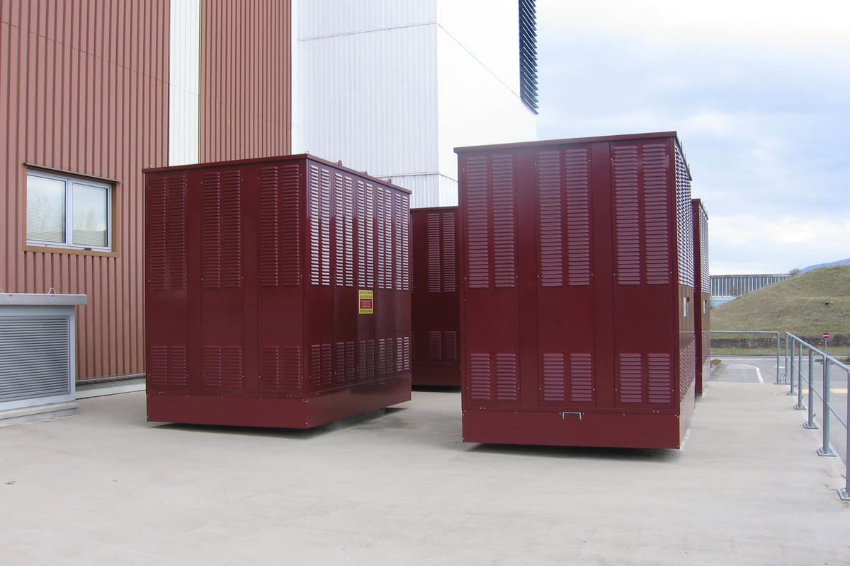 Two maroon ventilated transformer enclosures on a paved area beside a computer centre building