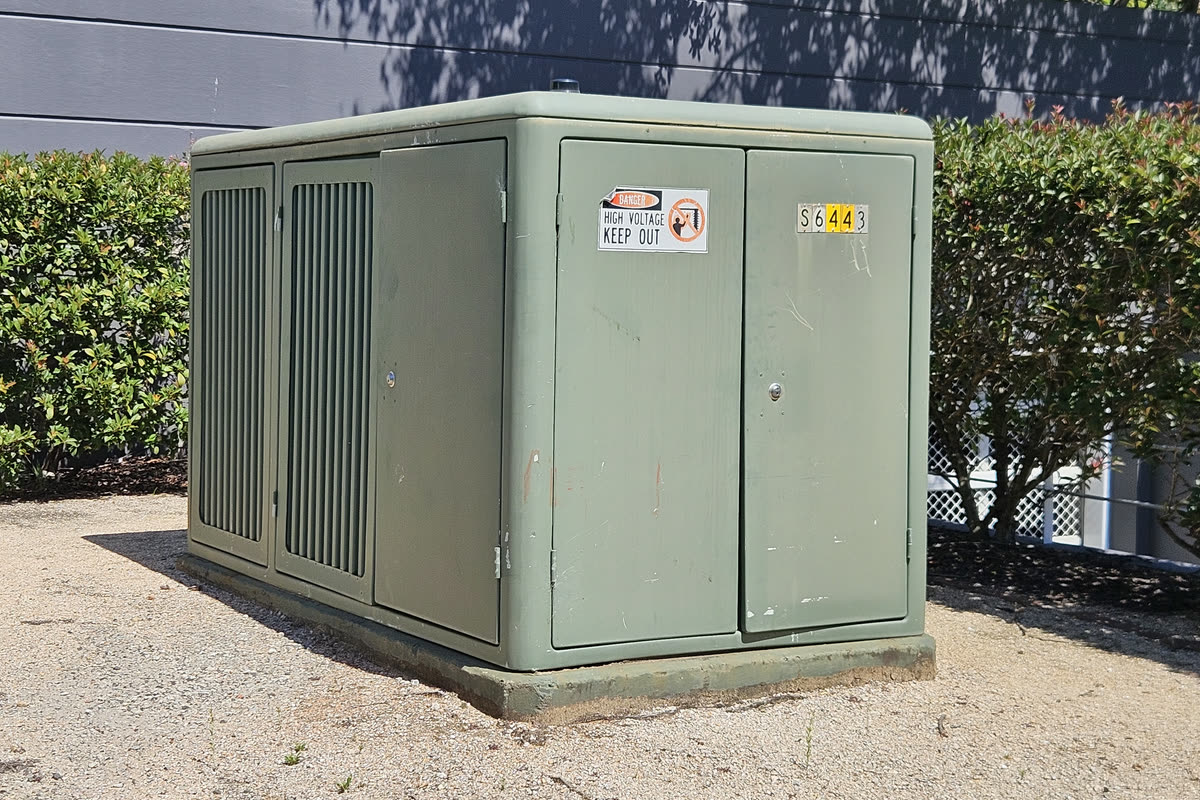 Green ground mounted substation enclosure with ventilated doors on a concrete plinth, between hedges