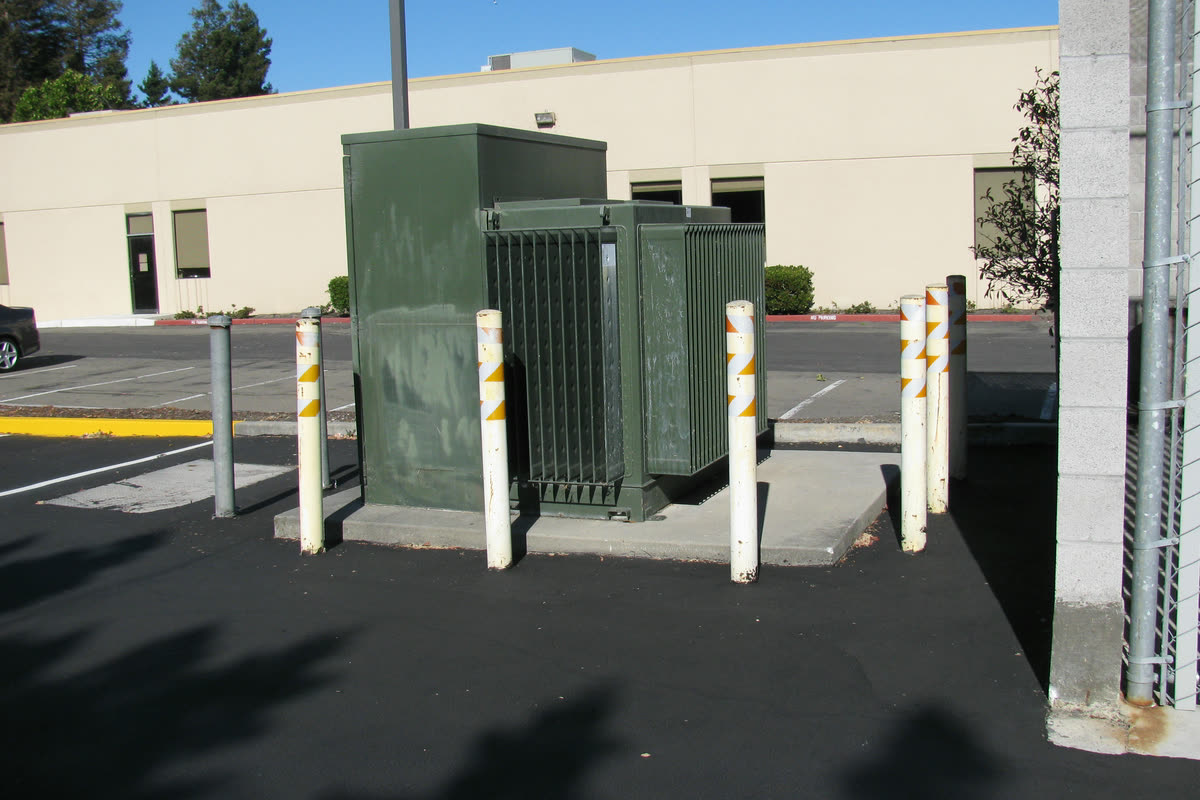 Green pad mounted transformer with cooling radiators on a concrete pad, protected by steel bollards outside a building