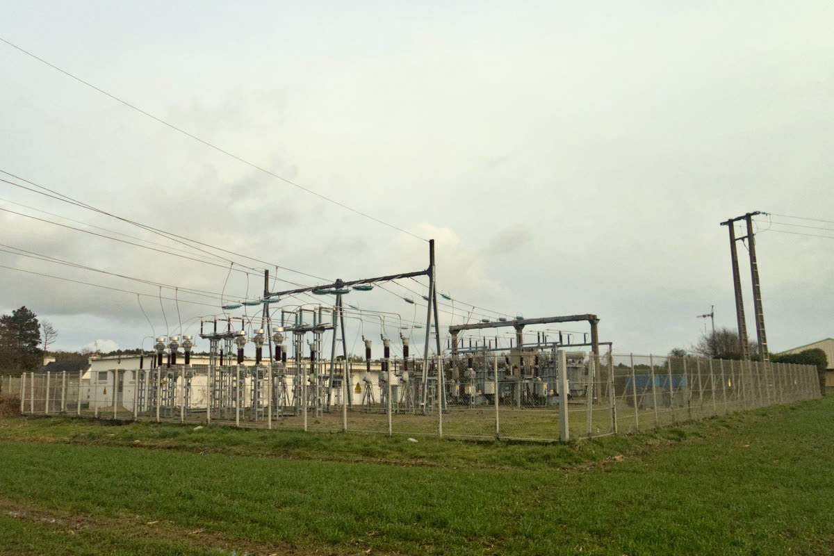 Outdoor high voltage substation with gantries and equipment behind a fence, seen across a grass field under an overcast sky